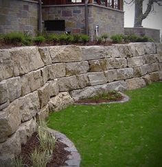 a stone wall in front of a house with green grass and bushes on the side