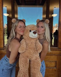 two beautiful women standing next to each other holding a large teddy bear