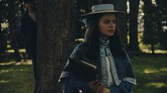 a woman wearing a hat and holding a book in her hands while standing next to a tree