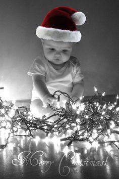 a black and white photo of a baby wearing a santa hat sitting in front of christmas lights