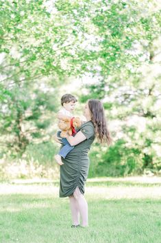 a woman holding a child in her arms while standing on the grass with trees in the background