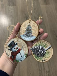 hand holding three christmas ornaments in front of a wood flooring background with a tree and snowman