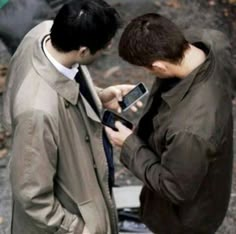 two men standing next to each other looking at their cell phones and texting on them