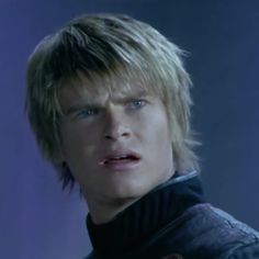 a young man with blonde hair and blue eyes wearing a black outfit looking at the camera