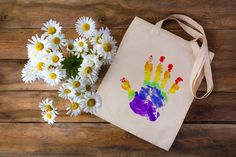 a tote bag sitting next to some daisies on a wooden table with a hand print