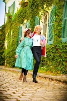 two women walking down a cobblestone road in front of green shuttered windows