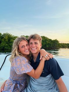 two people are sitting on a boat in the water and smiling at the camera, with trees in the background