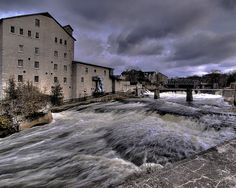 a river running through a small town next to a tall white brick building with a bridge in the background