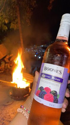 someone holding up a bottle of booze next to a campfire with fire in the background