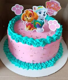 a pink and blue birthday cake with paw patrol decorations on it's top, sitting in a cardboard box