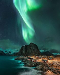 the northern lights shine brightly in the night sky above houses on rocky shore with water and snow covered mountains