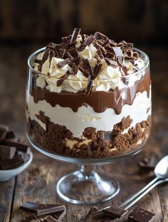 a dessert with chocolate and whipped cream in a glass dish next to spoons on a wooden table