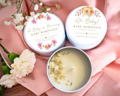 two small tins of body butter sitting on top of a pink cloth next to flowers