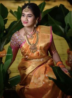 a woman in an orange and gold sari sitting on a bench with green leaves