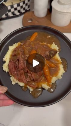 a person holding a plate with meat and vegetables on mashed potatoes in gravy