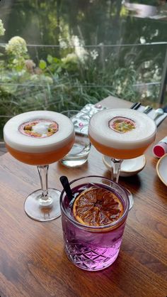 three glasses filled with drinks sitting on top of a wooden table next to plates and utensils