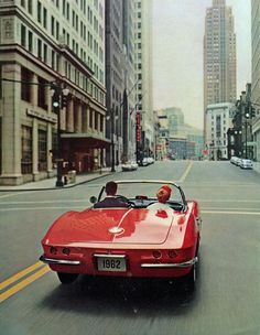 a red sports car driving down a city street with tall buildings in the back ground