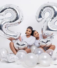 two women sitting in front of balloons with the number twenty three on them, surrounded by white and silver balloons