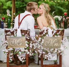 a bride and groom kissing in front of an outdoor table decorated with flowers, greenery and burlap signs