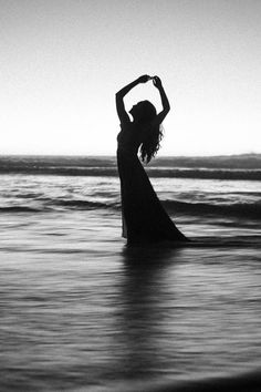 a woman standing in the ocean with her arms up and hands behind her head, wearing a long dress