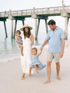 a man and woman holding hands while walking with two children on the beach next to a pier