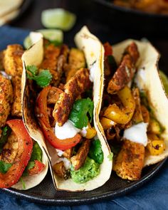 three tacos with chicken, peppers and lettuce on a black plate next to other food items