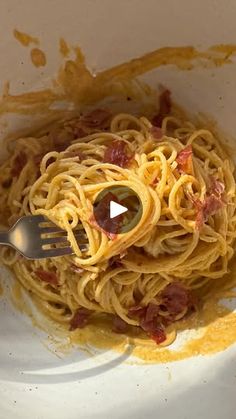 a white bowl filled with spaghetti and bacon on top of a table next to a fork