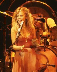 a woman in a pink dress holding a microphone and standing next to a drum set