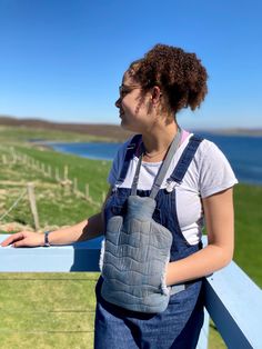 a woman wearing overalls and carrying a bag on her shoulder, looking at the ocean