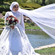 a woman in a white wedding dress holding a bouquet and veil over her head while standing next to a lake