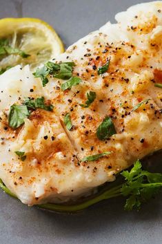 a fish fillet with herbs and lemon wedges on a gray surface next to a slice of lemon