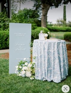 a table with a sign and flowers on it in front of some trees, grass and bushes