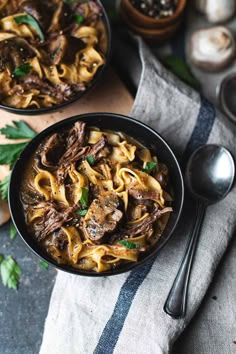 two bowls filled with beef and noodles on top of a table next to spoons