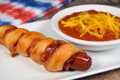 a hot dog wrapped in bacon next to a bowl of chili and cheese on a white plate