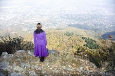 a woman in a purple dress standing on top of a hill looking at the valley below