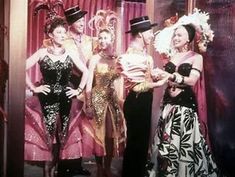 four women in dresses and hats posing for a photo on the set of an old fashion show