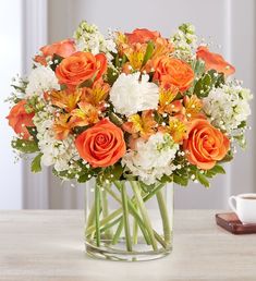 an orange and white flower arrangement in a glass vase on a table next to a coffee cup