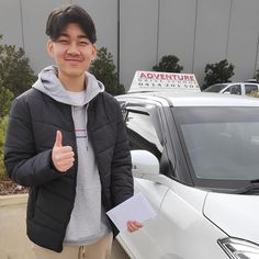 a young man standing in front of a white car giving the thumbs up sign with his hand