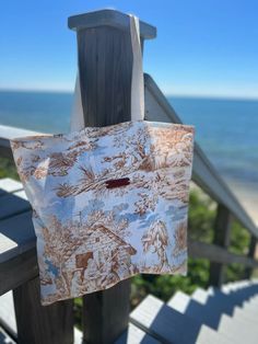 a brown and white tote bag hanging from a wooden railing near the ocean with trees