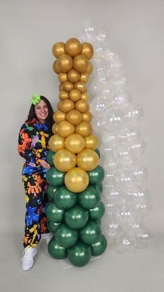 a woman standing next to a tall column made out of gold and green balloons in front of a white wall