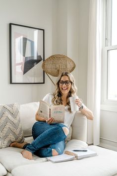 a woman sitting on top of a white couch holding an open book in her hands