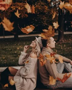 a man and woman sitting on a bench in the fall with leaves flying around them