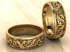 two gold wedding bands with engraved designs on them sitting on a wooden floor next to each other