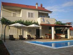 an empty swimming pool in front of a large house with patio furniture and umbrellas