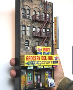 a person holding up a miniature model of a building in front of a phone booth