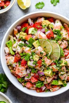 a white bowl filled with shrimp, avocado and tomato salad next to lime wedges