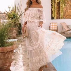 a woman standing next to a pool wearing a white dress and straw hat with her hands in her pockets