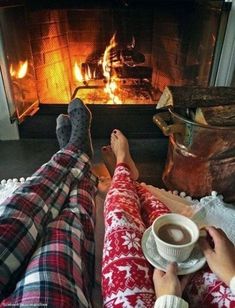 two people sitting in front of a fireplace with their feet up on a plate and holding a cup of coffee