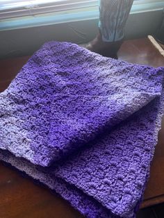 a purple crocheted blanket sitting on top of a wooden table next to a vase