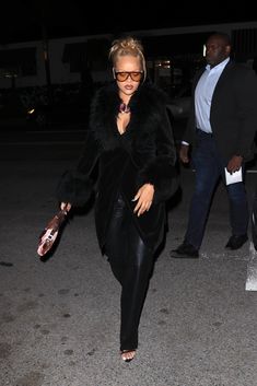 the woman is walking down the street in her black outfit and fur coat, with sunglasses on